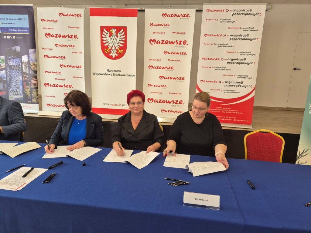 Trzy kobiety siedzą przy stole pokrytym niebieskim obrusem, podpisując dokumenty. W tle biało-czerwone bannery z napisem „Mazowsze” oraz herbem. Na stole leżą czarne długopisy i kartki.