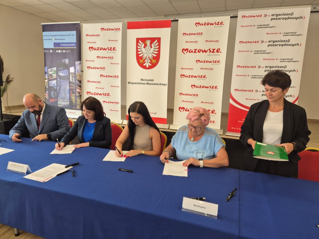 Ludzie podpisują dokumenty przy stole z niebieskim obrusem. Za nimi są plansze z logo i napisami Mazowsze. Pomieszczenie jest jasne, a ściany kremowe.