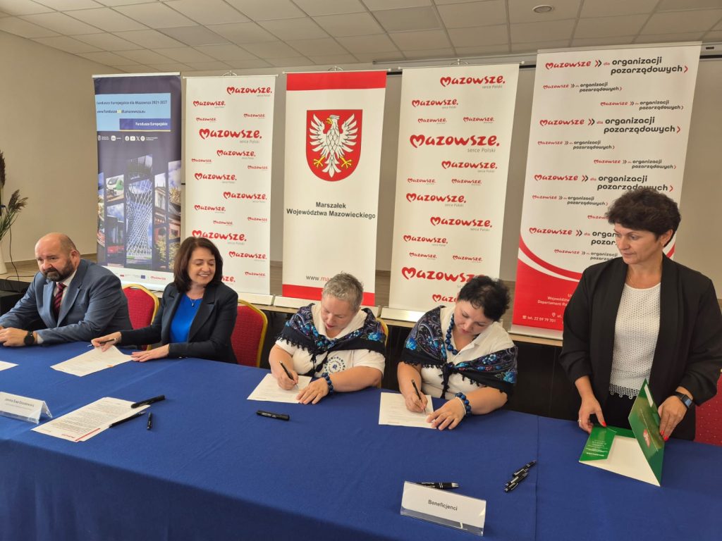 Na zdjęciu pięć osób przy stole z niebieskim obrusem, podpisujących dokumenty. W tle bannery z logo Mazowsza. Kilka z osób trzyma długopisy.