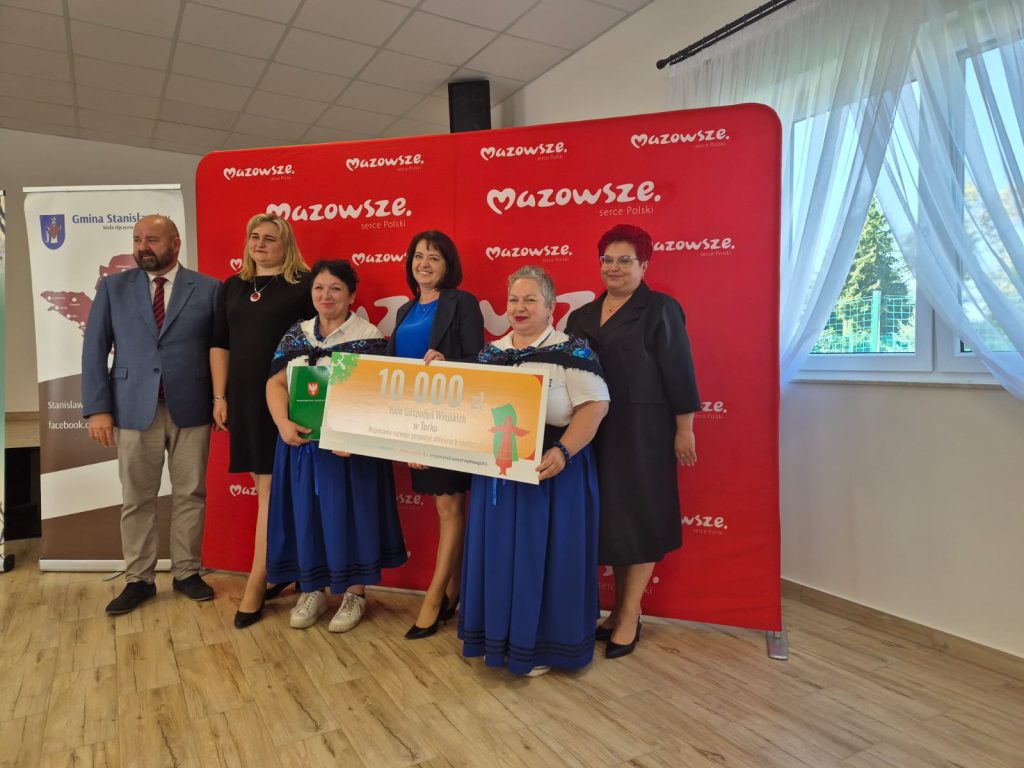 Grupa ludzi stoi na tle czerwonego baneru z napisem Mazowsze. Trzymają duży czek na 10 000 zł. Dwie kobiety mają niebieskie stroje ludowe.