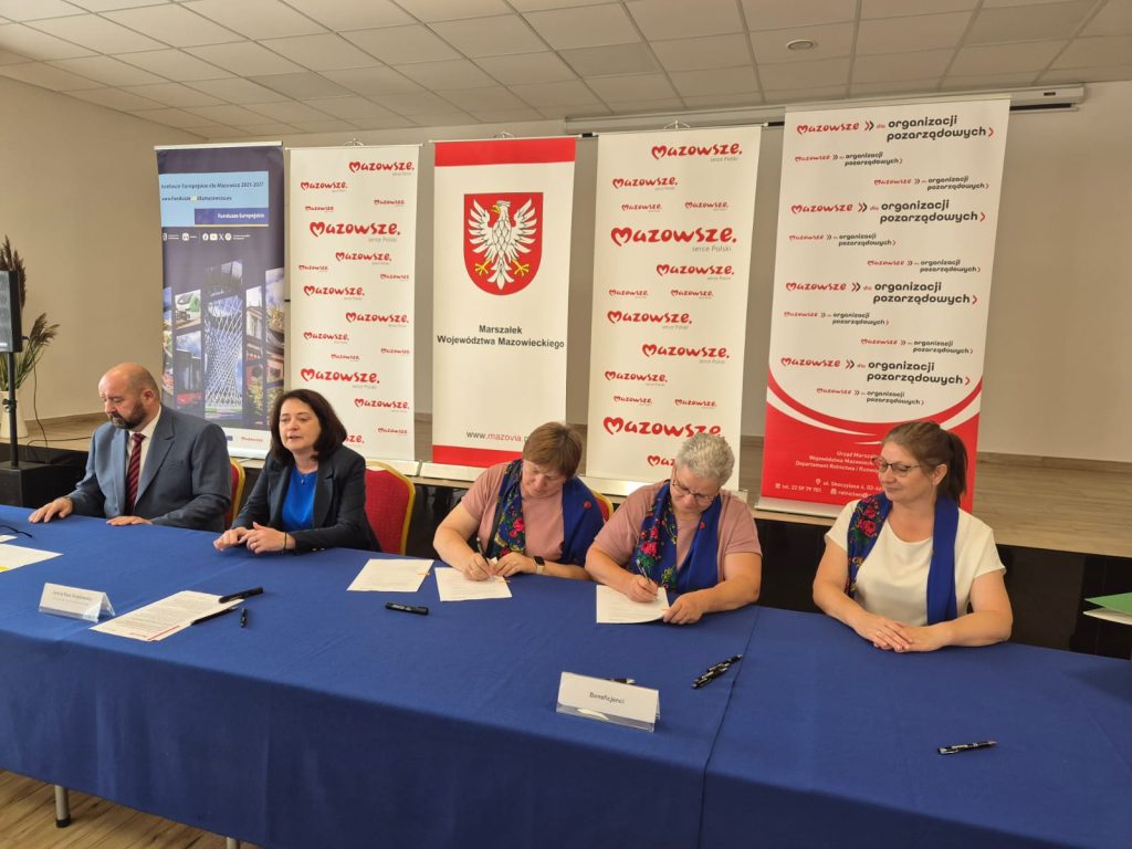 Na zdjęciu pięć osób siedzi przy stole z niebieskim obrusem. W tle wiszą banery z logo i napisami Mazowsze. Osoby podpisują dokumenty w jasnym pomieszczeniu.