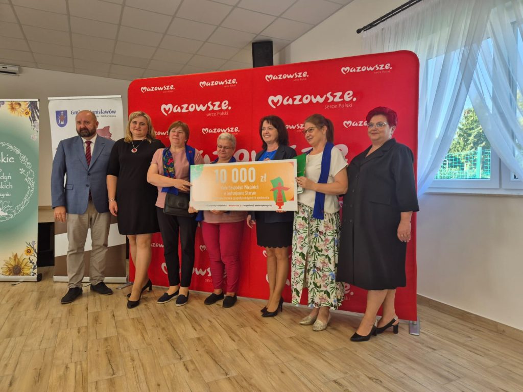 Grupa osób na tle czerwonego baneru z napisem Mazowsze, trzyma duży czek na jasnym tle drewnianej podłogi.