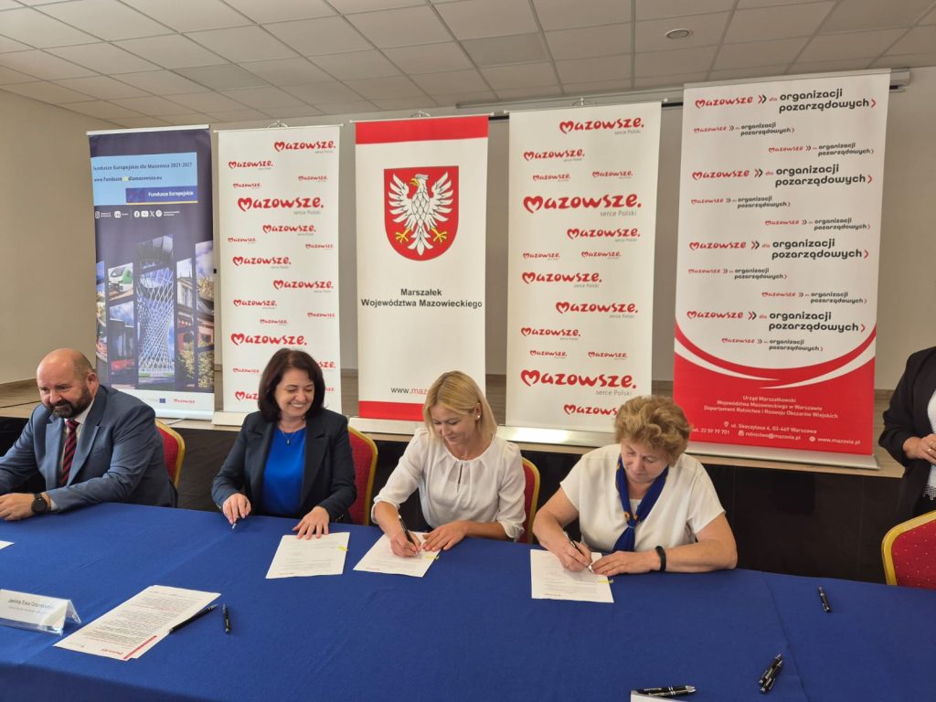 Cztery osoby siedzą przy stole z niebieskim obrusem, podpisując dokumenty. W tle informacje o Mazowszu i banery z logo. Pomieszczenie ma jasne ściany i sufit.