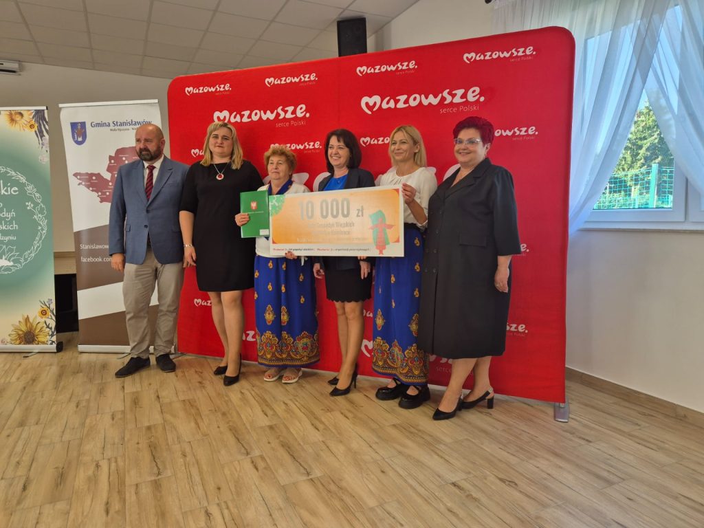 Grupa ludzi na tle czerwonego bannera Mazowsze trzyma duży czek na 10 000 zł. Osoby są elegancko ubrane. Podłoga jest drewniana, a w tle białe zasłony.