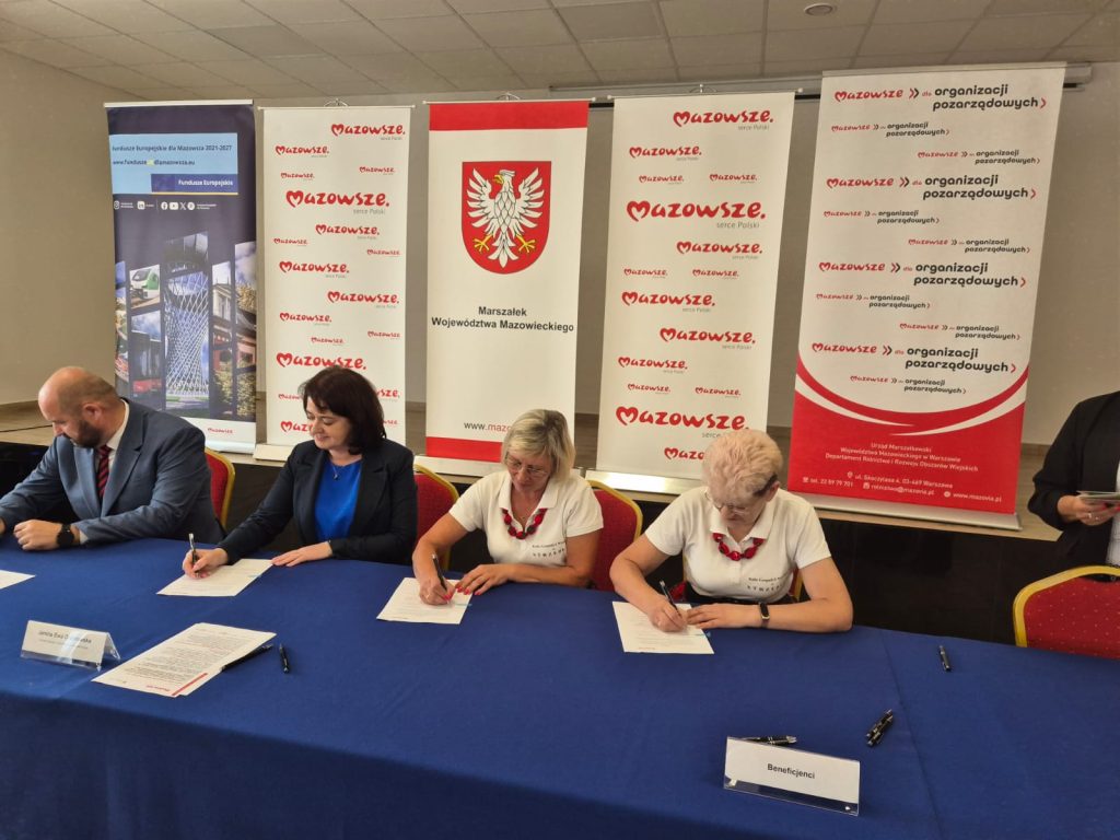 Trzy osoby siedzą przy stole z niebieskim obrusem, podpisując dokumenty. Za nimi wiszą plakaty z logo i nazwą Mazowsze. Tło jest jasne, z czerwonymi i białymi akcentami.