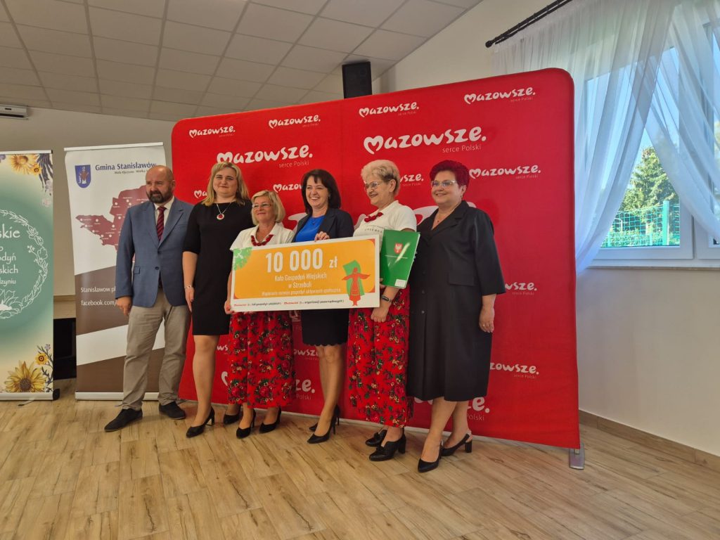 Sześć osób na tle czerwonego banera z białymi napisami. Trzymają duży czek na 10 000 złotych. Jasne, drewniane podłogi, białe zasłony po prawej.