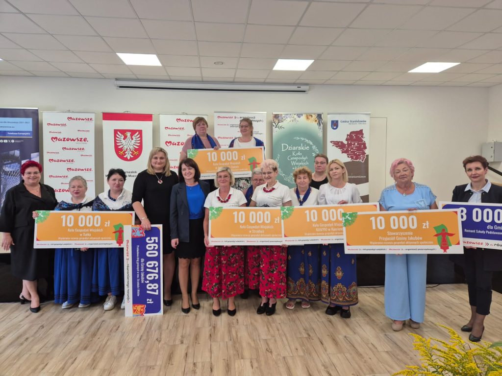 Grupa kobiet w strojach ludowych trzyma duże czeki. Jasne tło z plakatami i logotypami. Znajdują się w sali z drewnianą podłogą.