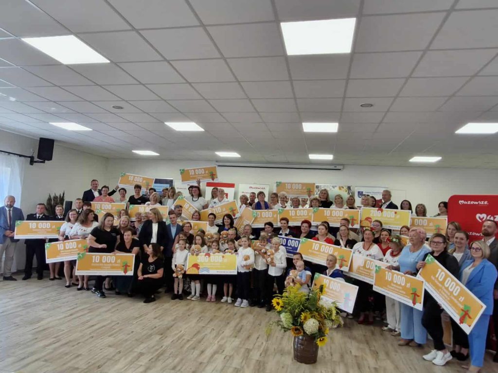 Grupa ludzi trzyma duże czeki z kwotą 10 000 zł na jasnym tle. Przód sali zdobią kwiaty. Wnętrze jest dobrze oświetlone.