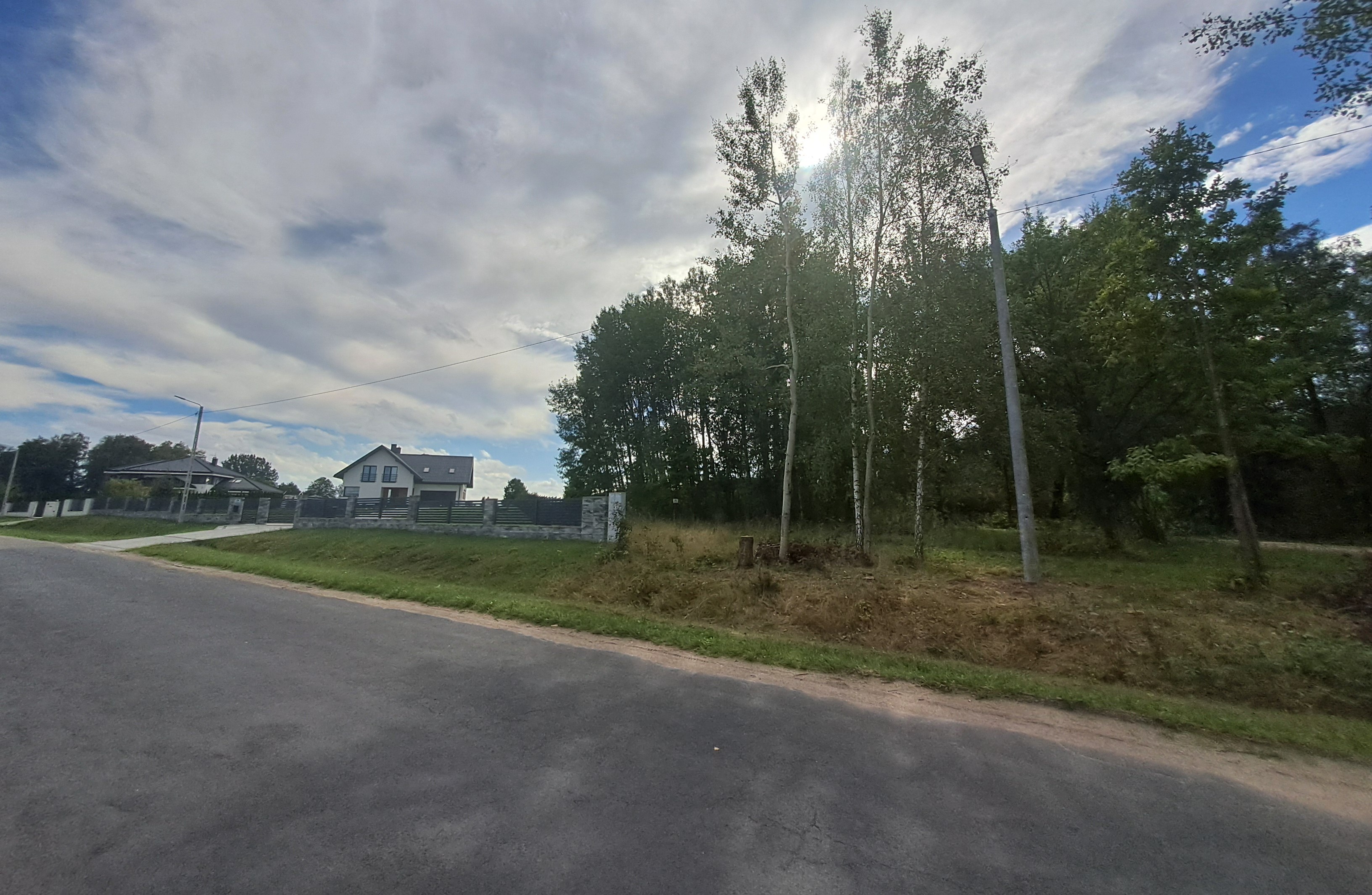 Droga asfaltowa z trawnikiem po bokach. Dom przy drodze, w tle białe chmury i niebieskie niebo. Po prawej drzewa.