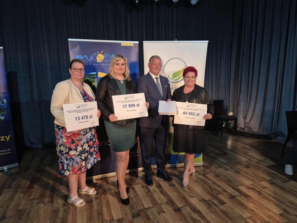 Na zdjęciu czworo ludzi na scenie trzymają tablice z kwotami pieniędzy. Kobiety w kolorowych ubraniach, mężczyzna w garniturze. Tło z ciemnymi zasłonami i banerami.
