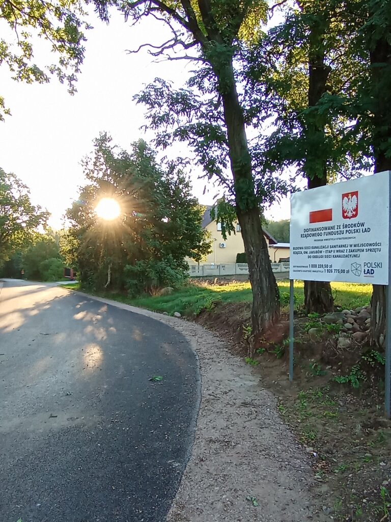 Droga biegnie przez zalesiony teren. Słońce świeci nisko na niebie. Po prawej stronie stoi tablica informacyjna.