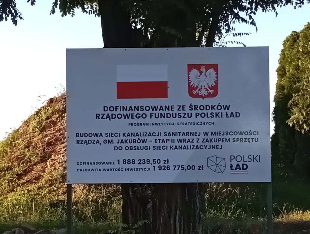 Tablica informacyjna o projekcie dofinansowanym przez Polski Ład; biały znak z flagą, godłem i tekstem, otoczony zielenią.