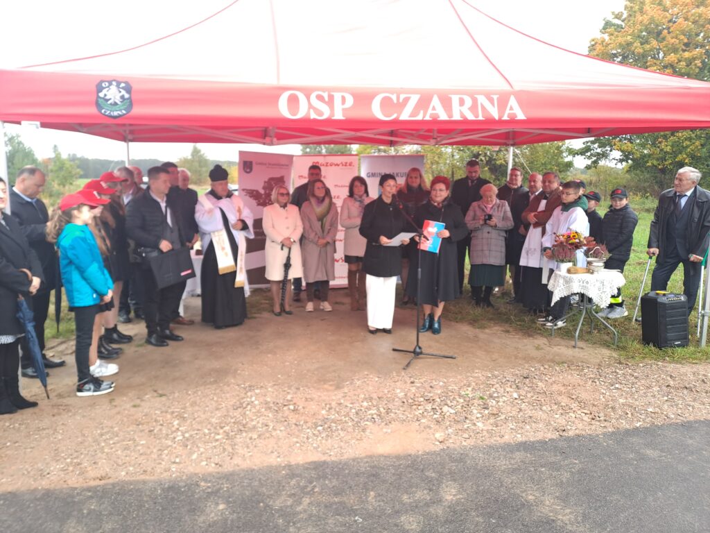 Grupa ludzi stoi pod czerwonym namiotem OSP Czarna. W tle drzewa i zielone otoczenie. Na stole znajduje się dekoracja z kwiatami.