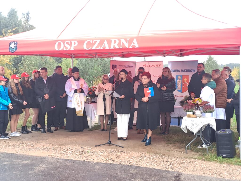 Grupa ludzi stoi pod czerwonym namiotem OSP Czarna. Wszyscy są ubrani formalnie, w tle są banery z napisami. Na stole po prawej stronie leżą kwiaty.