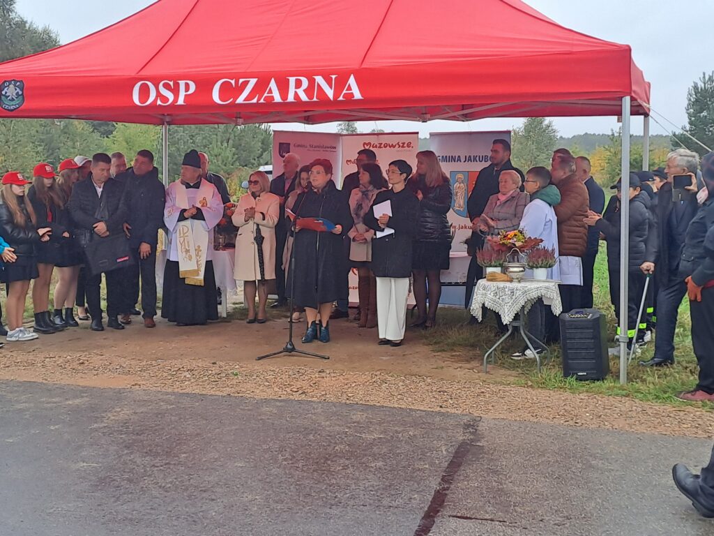 Grupa ludzi stoi pod czerwonym namiotem OSP Czarna na tle zieleni. Niektórzy trzymają mikrofony i notatki. Obok stołu z obrusem znajduje się ksiądz w białym ornacie.