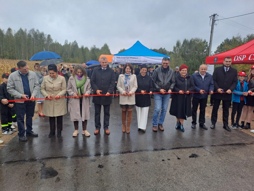 Grupa ludzi przecina czerwoną wstęgę na asfaltowej drodze. W tle namioty: niebieski i czerwony, oraz drzewa. Tło jest pochmurne i deszczowe.