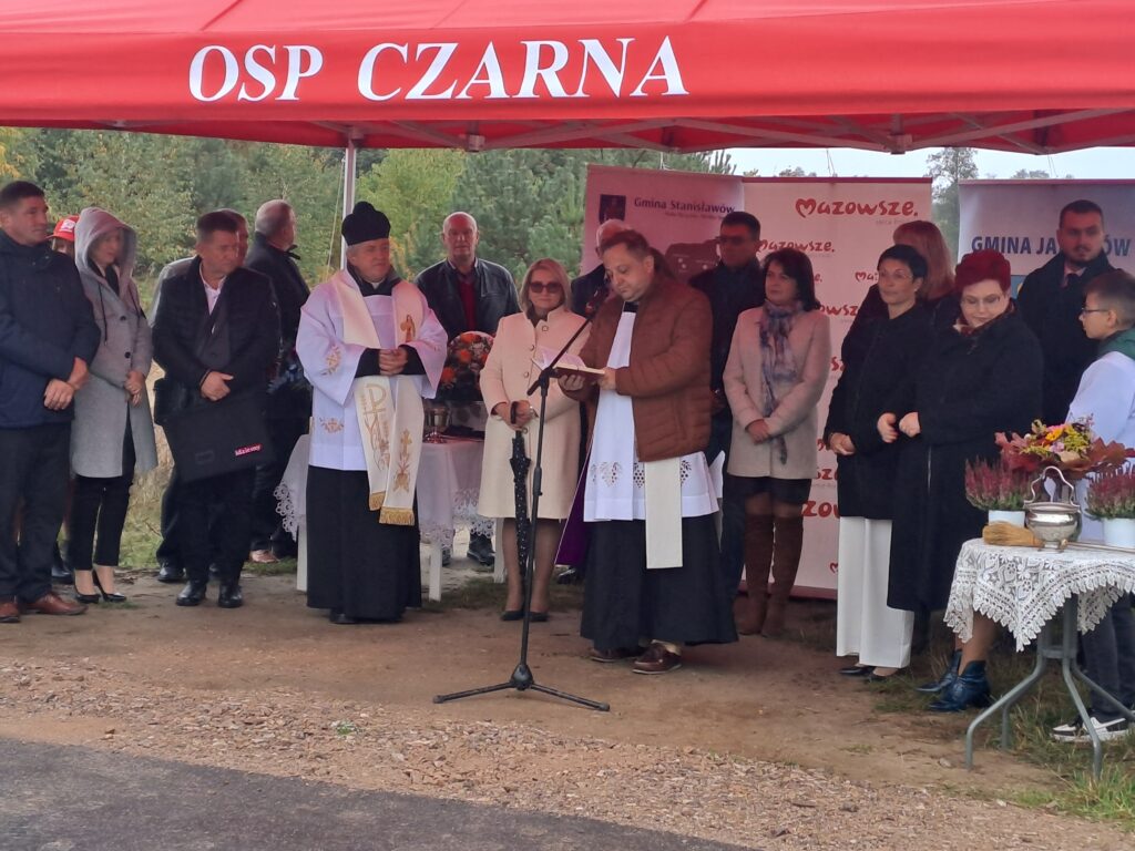 Ludzie zgromadzeni pod czerwonym namiotem OSP. Mężczyzna w szacie religijnej przemawia. Z tyłu zielone drzewa, po bokach bannery.