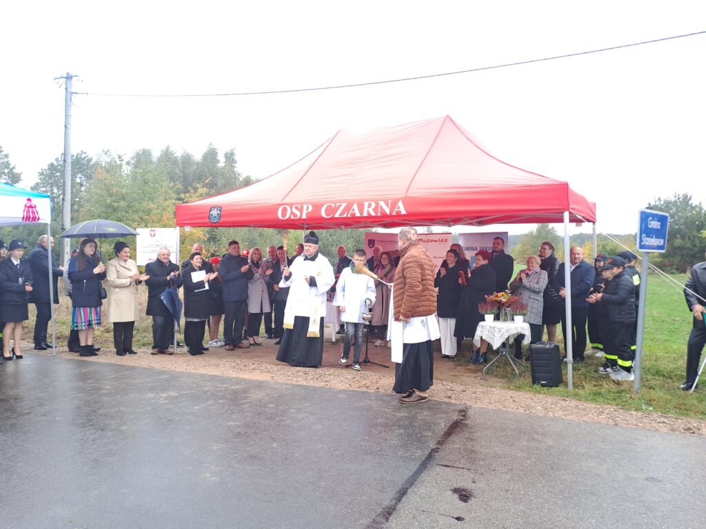 Grupa ludzi stoi pod czerwonym namiotem z napisem OSP Czarna. Na pierwszym planie duchowny w szatach liturgicznych. W tle zielone drzewa i znak Gmina Staszów.