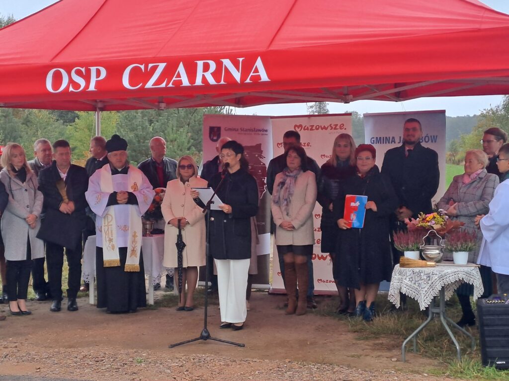 Grupa ludzi stoi pod czerwonym namiotem z napisem OSP CZARNA. Na pierwszym planie kobieta przemawia przez mikrofon, obok niej stół z kwiatami. Tło stanowi zielona roślinność i banery informacyjne.