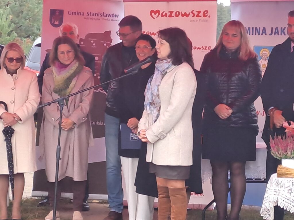 Grupa ludzi stojąca na scenie, kobieta przemawia do mikrofonu. W tle banery z logo Mazowsze, kolory dominujące to szarości i czerwienie. Widać też zieleń drzew w tle.