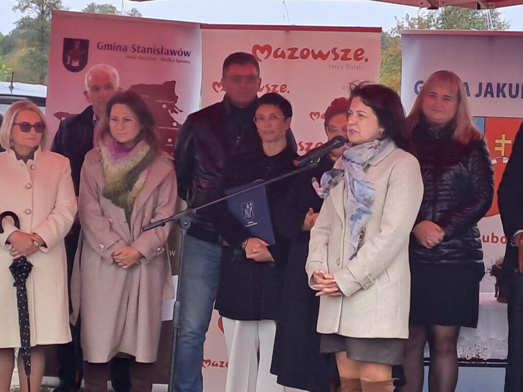 Na zdjęciu widać grupę osób w kurtkach i płaszczach na tle banerów Gmina Stanisławów i Mazowsze. Kobieta z szalikiem mówi do mikrofonu. Tło jest jasne, z elementami natury na górze.