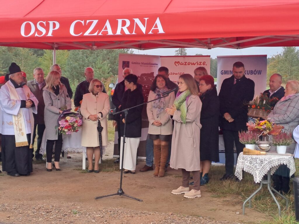 Grupa osób stoi pod czerwonym namiotem z napisem OSP CZARNA. Tło to zielone drzewa i plakaty promocyjne. Uczestnicy są elegancko ubrani, niektórzy trzymają kwiaty.