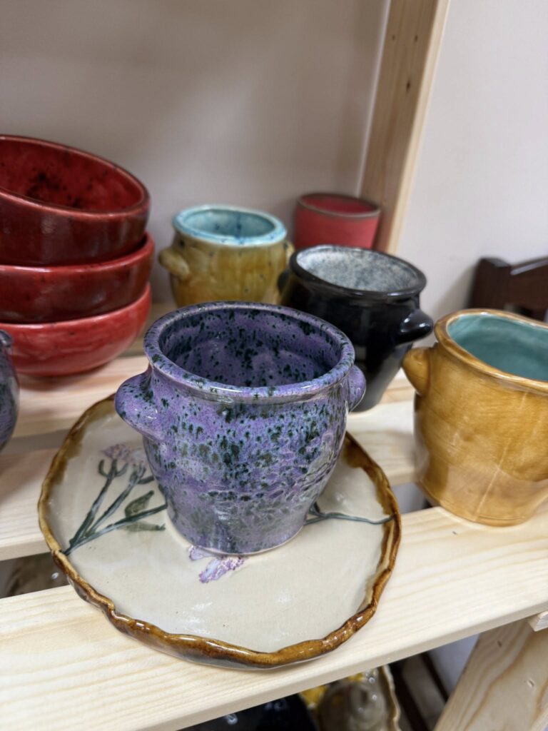 Na drewnianej półce stoją kolorowe ceramiczne kubki i talerz. Dominują barwy fioletu, żółci, czerwieni i niebieskiego. Talerz ozdobiony roślinnym wzorem.