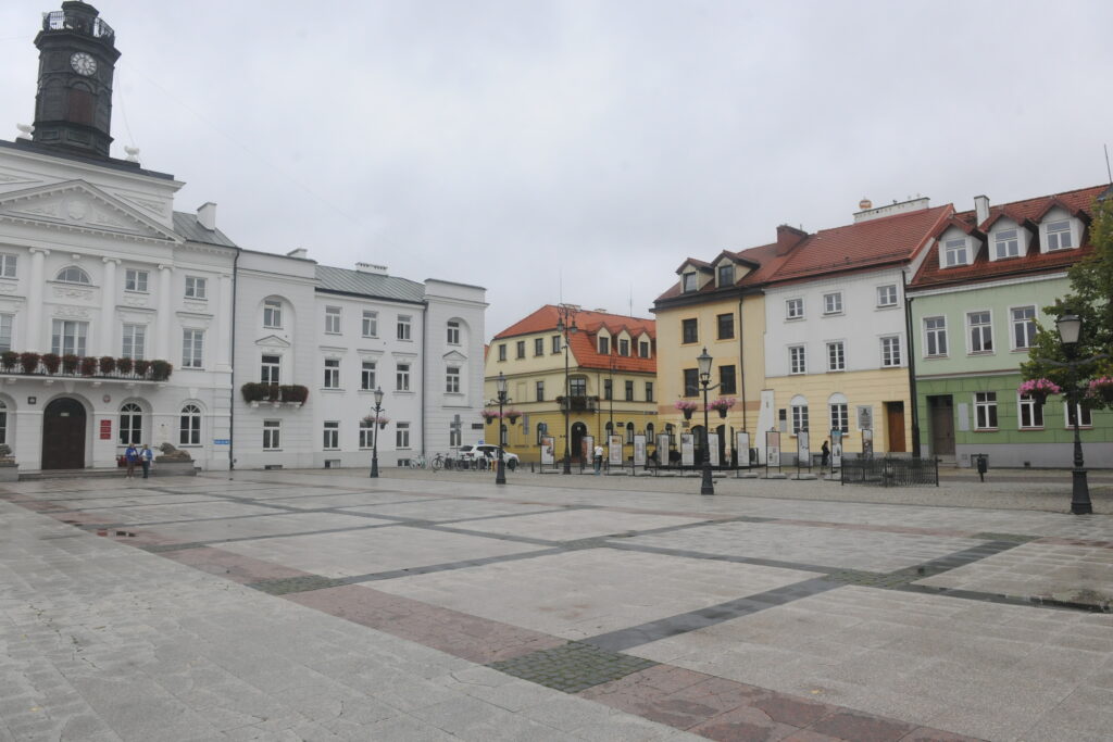 Rynek z zabytkowymi kamienicami w różnych kolorach: białym, żółtym, zielonym. Tło o pochmurnym niebie. Centralny plac jest pusty z nielicznymi dekoracjami.