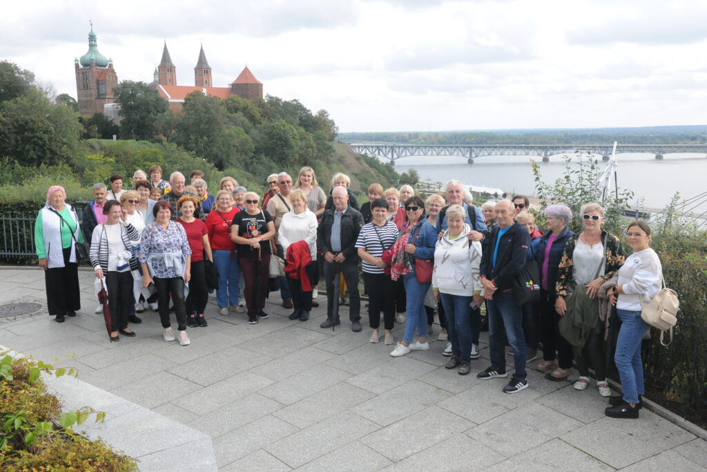 Grupa ludzi pozująca na tarasie z widokiem na rzekę. W tle most i zabytkowy budynek w otoczeniu zieleni. Niebo częściowo zachmurzone.
