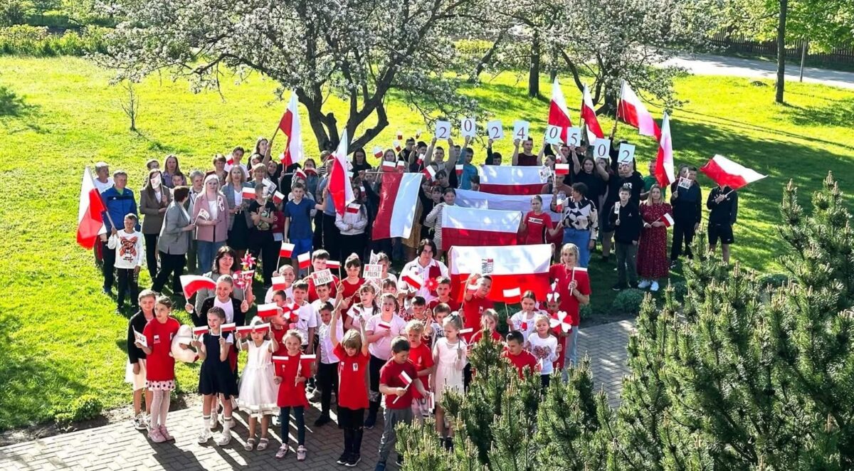 Grupa ludzi z flagami Polski pozuje na zielonym tle z drzewami. Ubrani są w czerwono-białe stroje. Trzymają także datę napisaną na kartkach.
