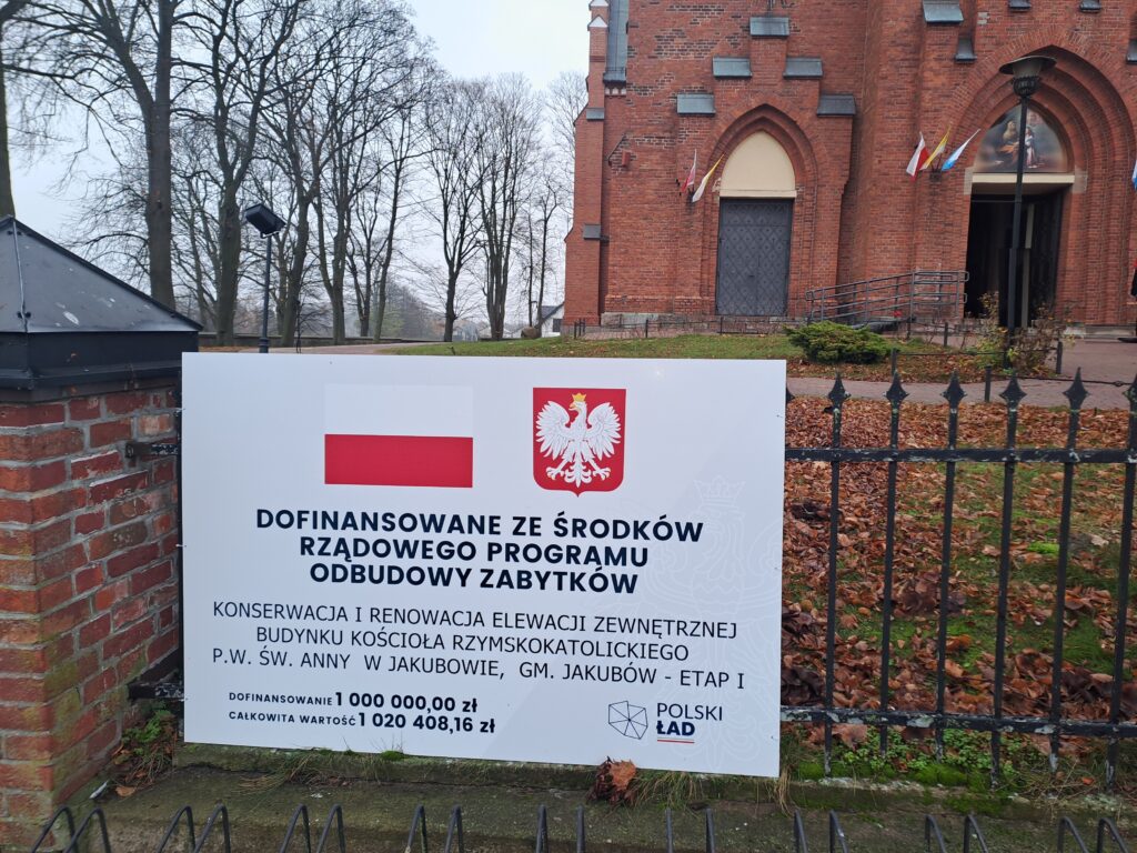 Na pierwszym planie widoczna tablica informacyjna z flagą Polski. W tle ceglany kościół i drzewa bez liści. Liście opadłe na ziemię, jesienna sceneria.