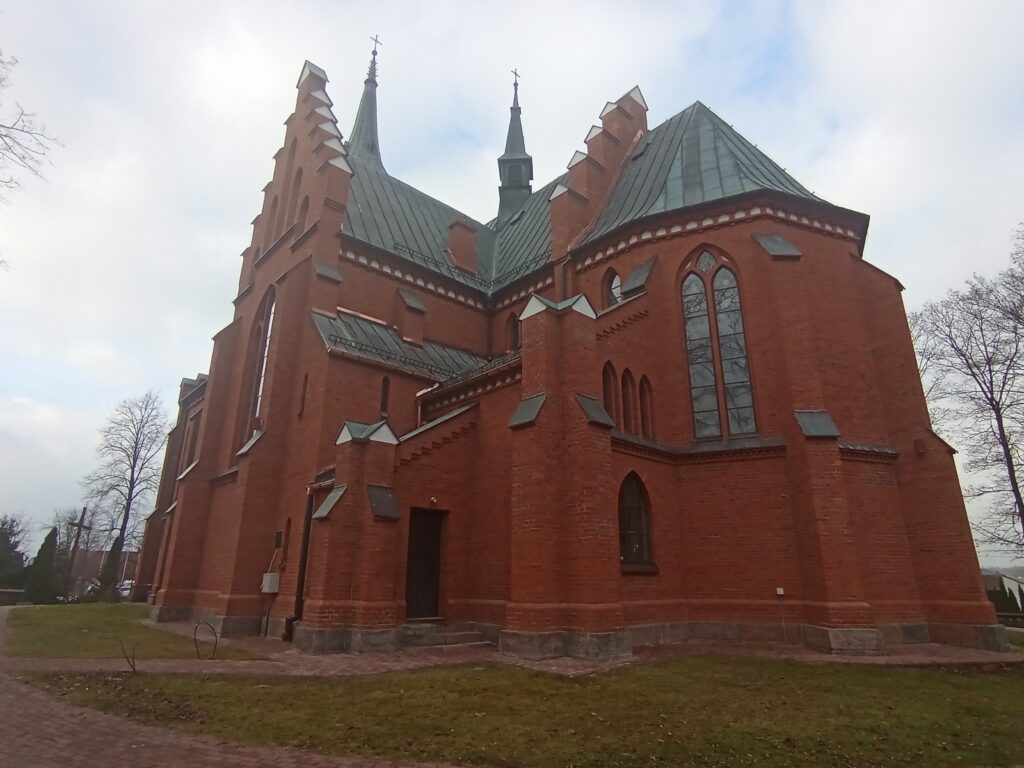 Ceglana gotycka świątynia z zielonym dachem i witrażowymi oknami. Tło to pochmurne niebo i kilka drzew. Brązowa brukowana ścieżka wokół kościoła.