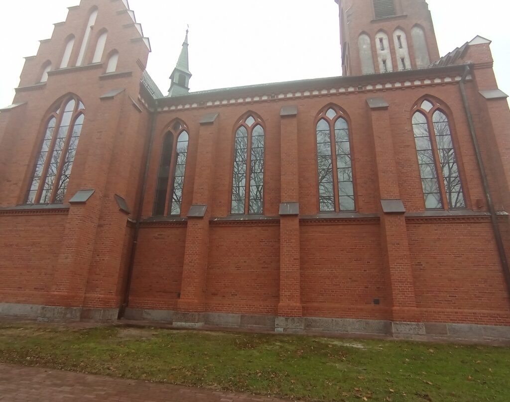 Ceglany kościół z wysokimi, strzelistymi oknami, na tle szarego nieba. Po lewej stronie widoczny fragment wieży. Trawa i brukowana ścieżka u podstawy budynku.