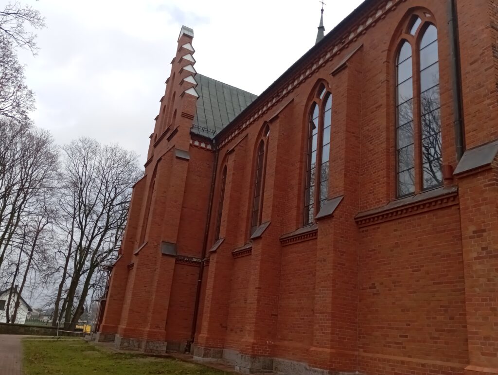 Ceglany kościół z dużymi, łukowatymi oknami na tle bezlistnych drzew. Budynek jest czerwony, a niebo szare i pochmurne. Drewniany dach zdobią ozdobne detale.