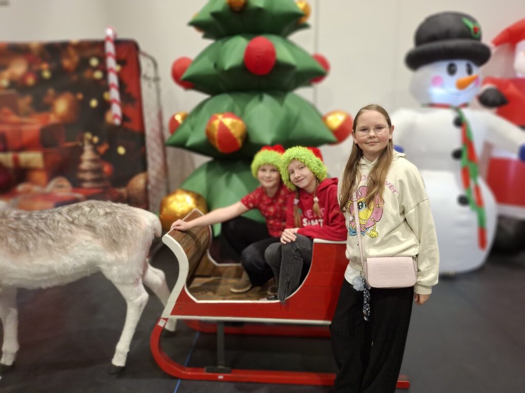 Na zdjęciu są trzy uśmiechnięte dzieci w scenerii świątecznej. W tle wielka zielona choinka i bałwan. Obok widoczny jest renifer i czerwone sanie.