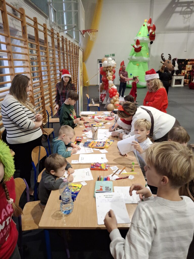 Grupa dzieci przy stole z kolorowankami i przyborami. W tle dmuchana choinka i ozdoby. Otoczenie sali gimnastycznej.