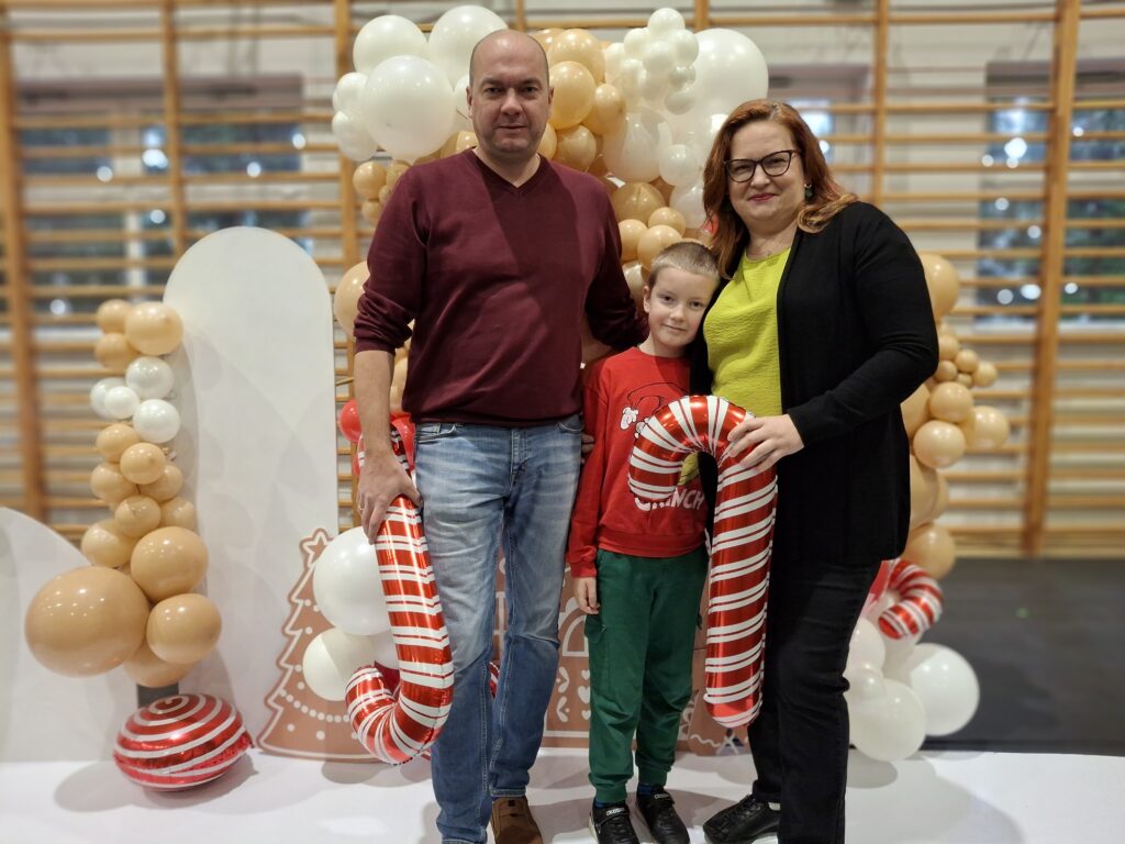 Trzy osoby stoją przed dekoracją z beżowymi i białymi balonami. Dziecko trzyma cukrową laskę w biało-czerwone pasy. Tło z jasnym drewnianym wzorem.