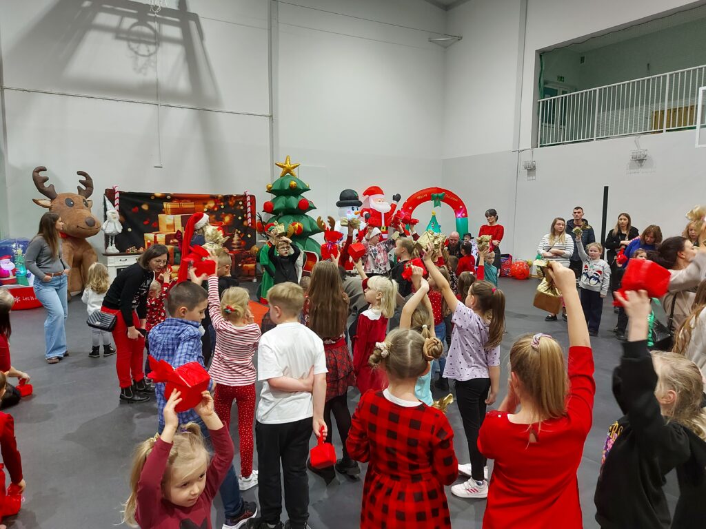 Dzieci bawią się na świątecznej imprezie w sali. Dekoracje przedstawiają renifera, choinkę i Mikołaja. Wesoła atmosfera, dominują czerwone i zielone kolory.
