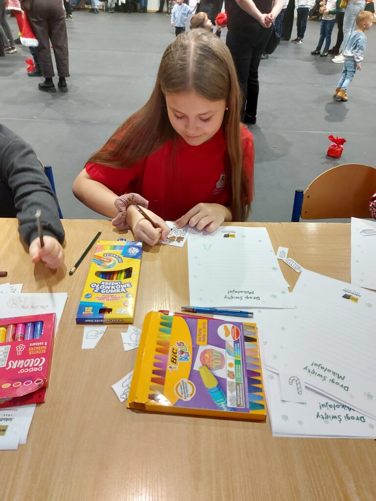 Dziewczynka w czerwonej koszulce rysuje przy stole pełnym kredek. Tło jest szare, z ludźmi w oddali. Na stole leżą kolorowe opakowania kredek i kartki.