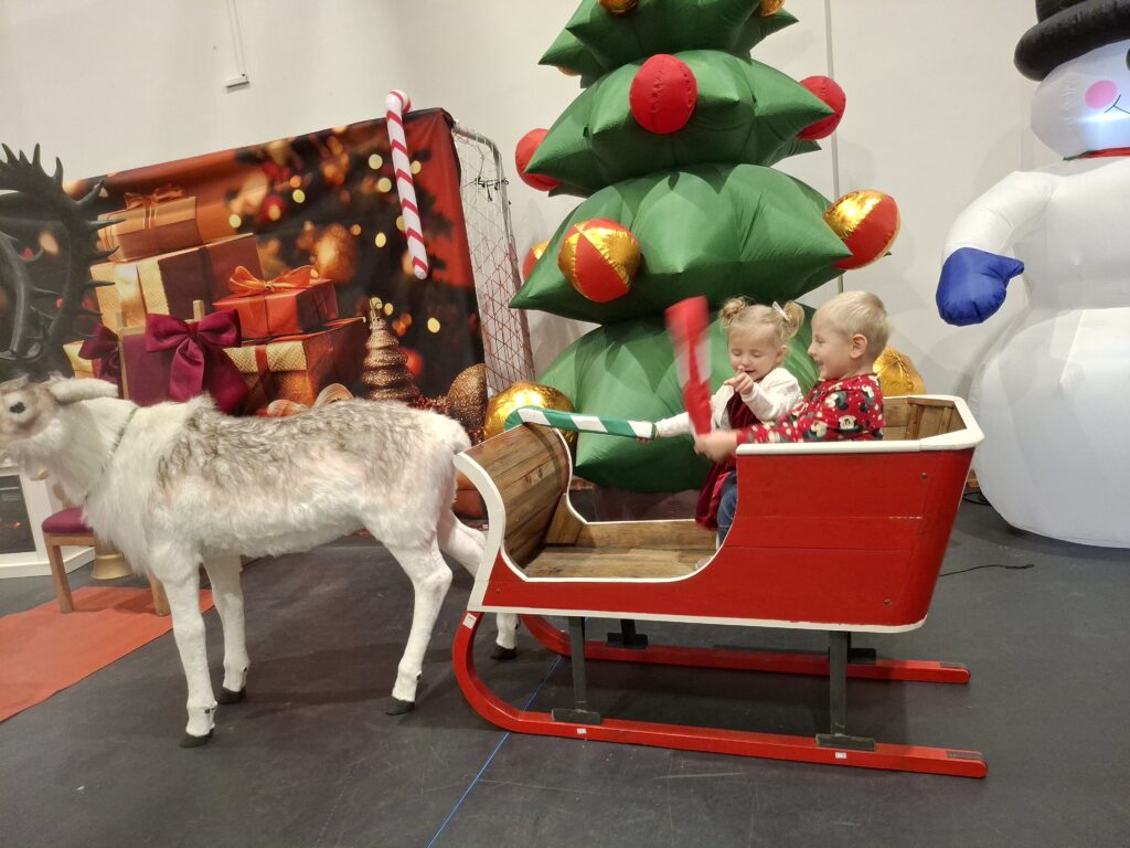 Dwójka dzieci w czerwonych sankach z reniferem. Tło to świąteczne dekoracje z zieloną choinką. Obok stoi biały bałwan.