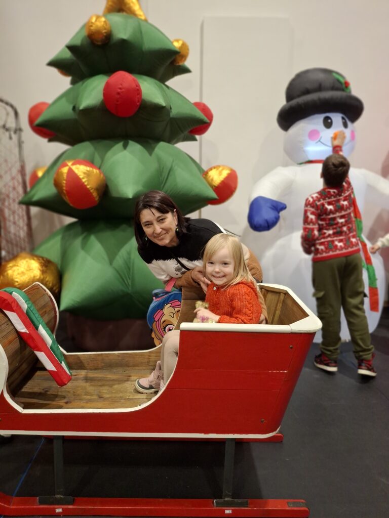 Na zdjęciu jest dziecko i osoba dorosła w czerwonych saniach. Obok stoi duża, dekoraowana choinka i bałwan. Tło jest jasne.
