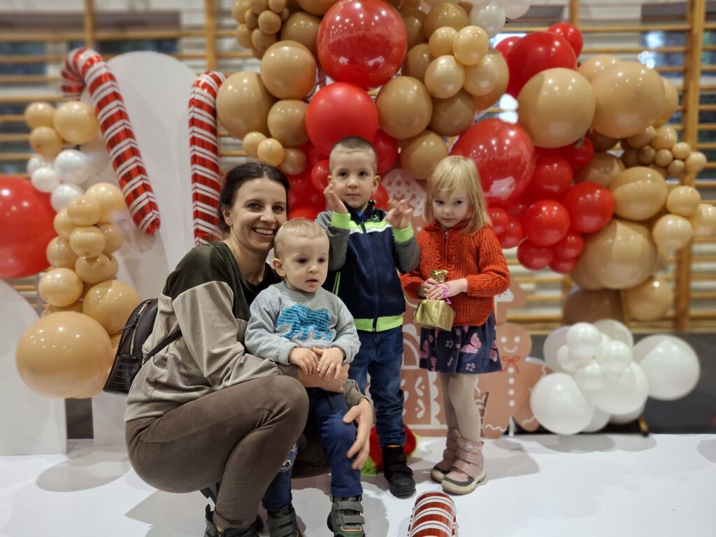 Na zdjęciu jest kobieta z trójką dzieci na tle balonowej dekoracji. Balony są w kolorach czerwonym, białym i brązowym. Tło zdobią także dekoracje w kształcie lasek cukrowych.