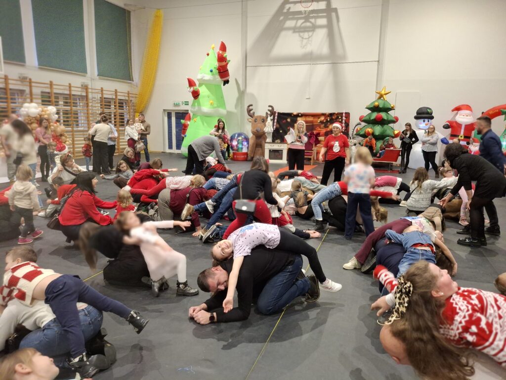 Na zdjęciu grupa dzieci i dorosłych bawi się w sali gimnastycznej. W tle widać kolorowe, dmuchane dekoracje świąteczne. Ubrania uczestników dominują w odcieniach czerwieni i bieli.