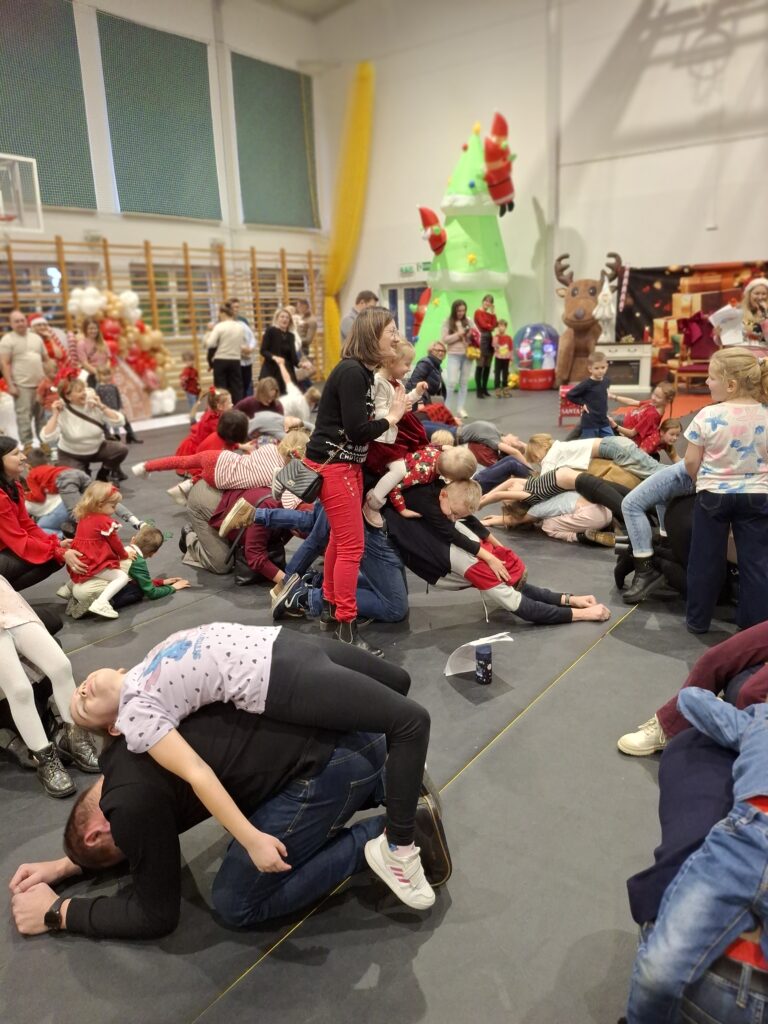 Na zdjęciu widać grupę ludzi w sali gimnastycznej. Widoczne są dekoracje świąteczne i nadmuchana choinka. Ubrani są w kolorowe stroje, głównie czerwono-białe.