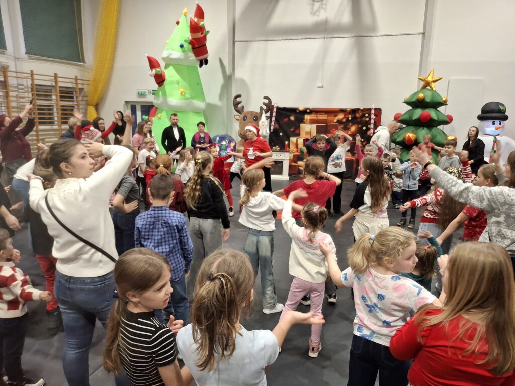 Grupa dzieci bawi się w sali gimnastycznej udekorowanej na święta. W tle są dmuchane choinki i bałwan. Ubrania uczestników są kolorowe, dominują czerwienie.