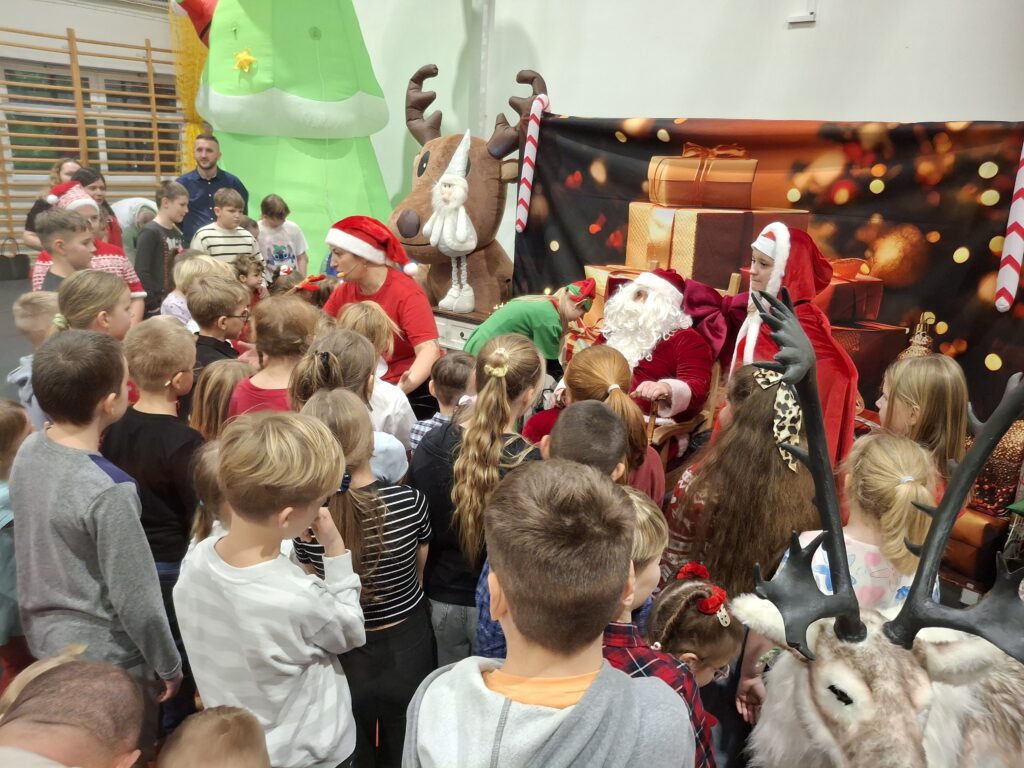 Grupa dzieci otacza Świętego Mikołaja w czerwonym stroju. W tle widać zielone dekoracje i ogromną pluszową postać renifera. Atmosfera jest świąteczna i kolorowa.