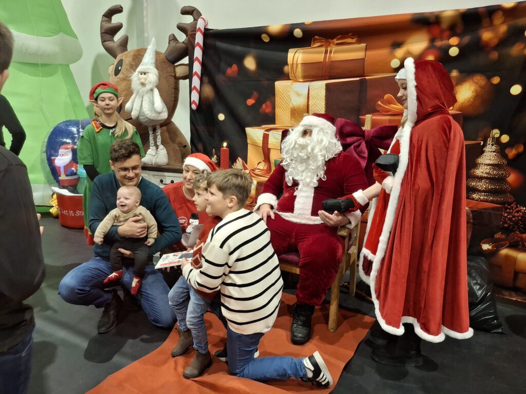 Grupa osób przy Mikołaju na tle prezentów. Dużo czerwieni i zieleni, atmosfera świąteczna. Renifer, elf, dzieci i dorośli wokół.
