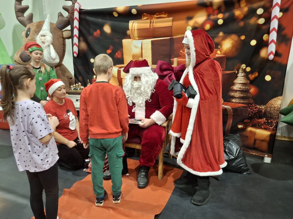 Święty Mikołaj z pomocnikami, dzieci stoją przed nim. Tło z prezentami i dekoracjami świątecznymi. Ciepłe, świąteczne kolory.