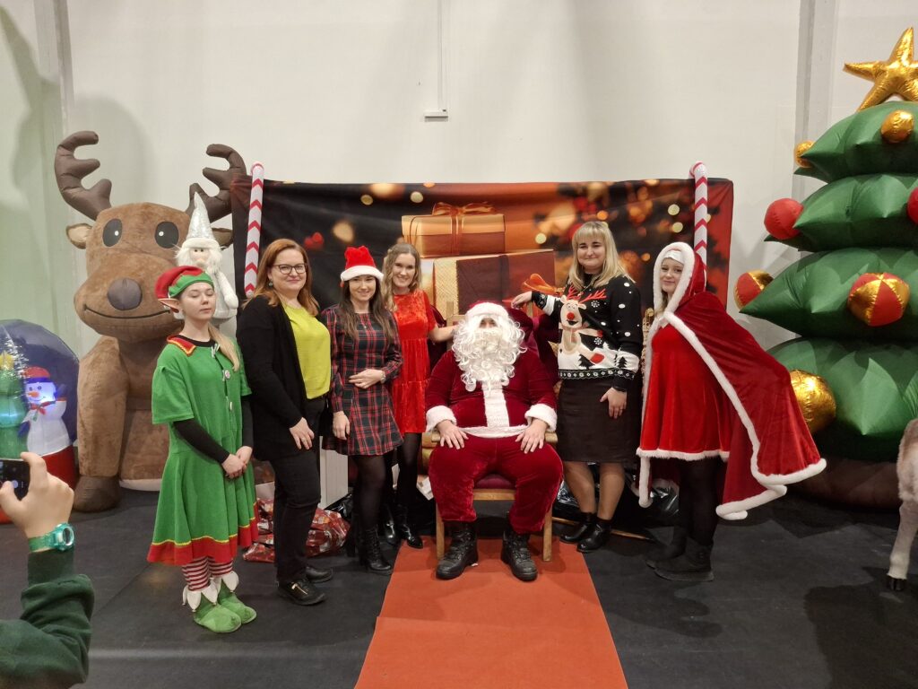 Grupa osób w świątecznych strojach, w tym Święty Mikołaj, stoi na tle z reniferem i choinką. Kolory: czerwony, zielony, biały. Tło ozdobione świątecznymi dekoracjami.