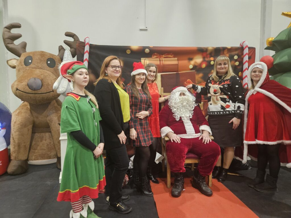 Święty Mikołaj w czerwonym stroju siedzi otoczony przez osoby w świątecznych kostiumach. Tło ozdobione reniferami i dekoracjami. Dominują kolory czerwony i zielony.